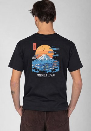 Mann von hinten, trägt ein schwarzes T-Shirt mit farbigem Motiv von Mount Fuji, Pagoden und orangefarbener Sonne, kombiniert mit brauner Hose.