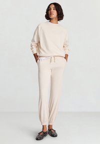 Felpa beige con maniche raglan e scollo rotondo; pantaloni coordinati con coulisse e polsini affusolati; scarpe nere con occhielli decorativi.