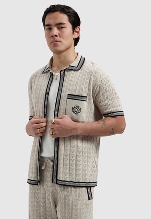 Jonge man past een open beige gebreid shirt met korte mouwen, zwarte bies en geborduurde zak aan, draagt bijpassende broek en gouden sieraden.