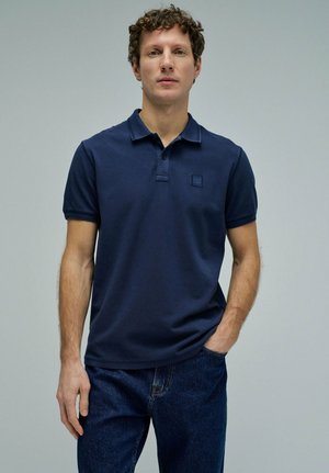 Jeune homme aux cheveux bouclés portant un polo bleu marine à manches courtes et un jean bleu foncé, debout devant un fond clair uni.