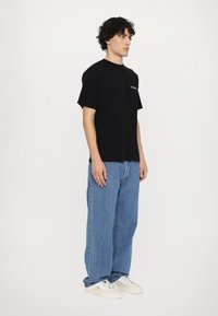 Svart bomullst-skjorte med korte ermer og rund hals, kombinert med løse blå denimjeans og hvite joggesko, stående på en ensfarget bakgrunn.