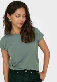 Camicia verde con una sottile texture a righe e maniche arrotolate, abbinata a pantaloni in corduroy verde scuro. Modella con orecchini d'oro.