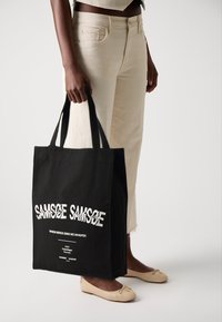 Black canvas tote bag with white text reading "SAND" and "WHEN BIRDS SING WE WHISPER." Sturdy handles, rectangular shape, minimalistic design.