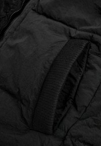 Details der schwarzen Pufferjacke mit einem strukturierten Stoff, einem gesteppten Design und einem auffälligen schwarzen Gummiband auf der rechten Seite.