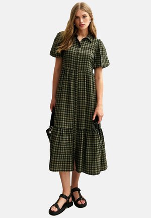 Robe midi en tartan vert avec des manches courtes bouffantes, un devant à boutons, une jupe à volants et une coupe ample, associée à des sandales noires et un sac à main.