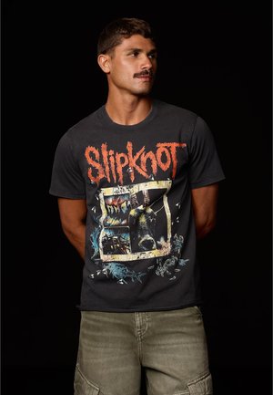 Lyhyttukkainen ja viiksin mies, jolla on musta Slipknot-yhtyeen t-paita ja oliivinväriset cargo-shortsit, seisoo mustaa taustaa vasten.