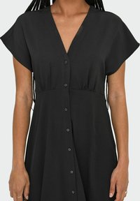 Robe noire à manches courtes, à col en V et avec des boutons sur le devant. Fabriquée dans un tissu léger, elle possède une taille froncée pour un ajustement décontracté.