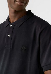 Polo shirt nero con tessuto testurizzato, chiusura a bottoni e un logo in gomma sottile sul lato sinistro del petto. I bottoni sono bianchi con centri neri.