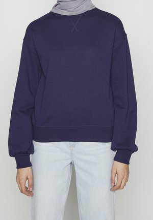 Person iført en marineblå sweatshirt, lyseblå jeans og et lysegråt hovedtørklæde, stående mod en ensfarvet baggrund.