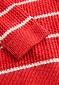 Roter gestrickter Pullover in der Nahaufnahme mit einem gerippten Bündchen, strukturiertem Muster und weißen horizontalen Streifen auf einem glatten Hintergrund.