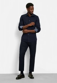 Marineblaues Hemd mit Knopfleiste und kontrastierenden hellblauen Manschetten, kombiniert mit dunkler Hose und schwarzen Schuhen, das eine körperbetonte Silhouette zeigt.