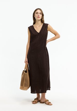 Frau in ärmellosem, dunkelbraunem Midi-Kleid mit V-Ausschnitt, die eine hellbraune, perforierte Tragetasche hält und hellbraune Sandalen trägt, vor weißem Hintergrund stehend.