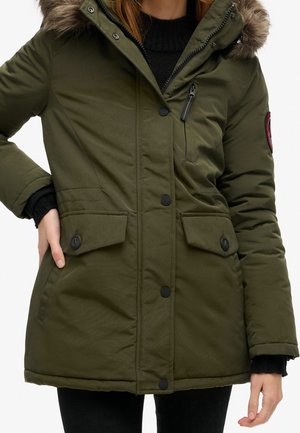 Kvinde iført en olivengrøn parka med pelskant på hætten, sort sweater og sorte bukser, hænderne hvilende nær lommerne.