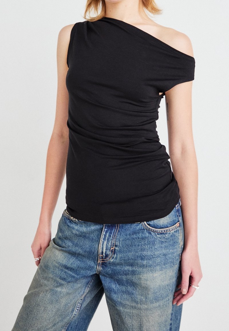 Femme portant un top noir asymétrique et un jean bleu ample, debout devant un fond clair uni.
