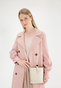 Lyserød stof trenchcoat med store sorte knapper, iført en lys beige crossbody taske med tekstureret overflade og fremtrædende håndtag.
