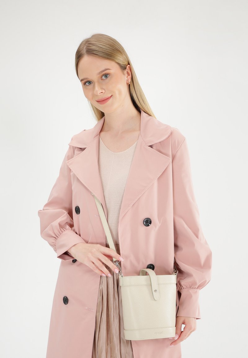 Lyserød stof trenchcoat med store sorte knapper, iført en lys beige crossbody taske med tekstureret overflade og fremtrædende håndtag.