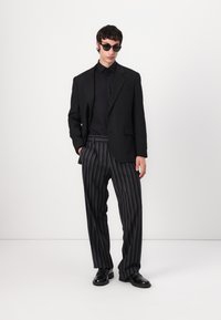Black blazer over a black shirt, paired with black and white striped trousers. Accessories include round black sunglasses and black shoes.