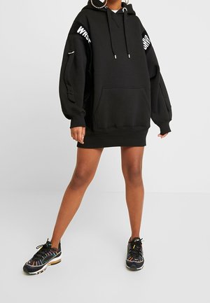 Osoba nosząca oversize'ową czarną sukienkę z kapturem z białym tekstem na rękawach oraz czarne sneakersy z kolorowymi paskami stoi na jednolitym tle.