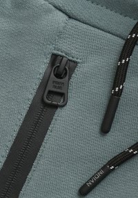 Ljusgrön sweatshirt-tyg med en texturerad yta. Har en svart dragkedja och snoddar med kontrasterande vita accenter.