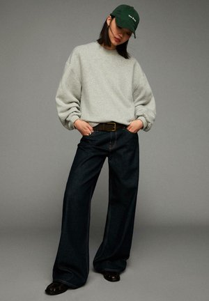 Sweat à capuche gris surdimensionné, jean large bleu foncé, casquette verte avec texte. Chaussures noires. Coupe décontractée avec des manches à revers et des coutures visibles.