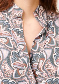 Blouse à motifs avec col, présentant un mélange de couleurs vert, rose et marron. Le design comprend des motifs spiraux et des détails complexes.