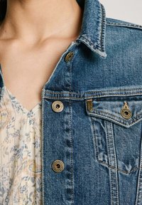 Chaqueta de denim con un lavado azul clásico, detalles de botones y un bolsillo en el pecho con etiqueta de logo. Tejido texturizado y diseño de cuello estructurado.