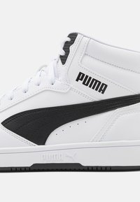 Biała sneakersy typu high-top Puma z czarnym paskiem, czarnym logo Puma z boku, białymi sznurówkami i wytłoczonym logo Puma na białej podeszwie.