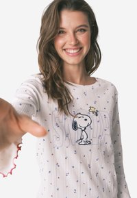Haut à manches longues à côtes blanches avec des motifs floraux multicolores, présentant un graphique de Snoopy et le texte "SNOOPY" en lettres audacieuses, bords des manches rouges à volants.
