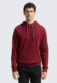 Felpa rossa con cappuccio, tasca frontale, cordini e tasca con zip sulla manica. Polsini e orlo a coste con una texture liscia.