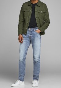 Olivgrön bomullsjacka med tryckknappar, svart t-shirt, ljusblå jeans med blekt finish och vita sneakers.