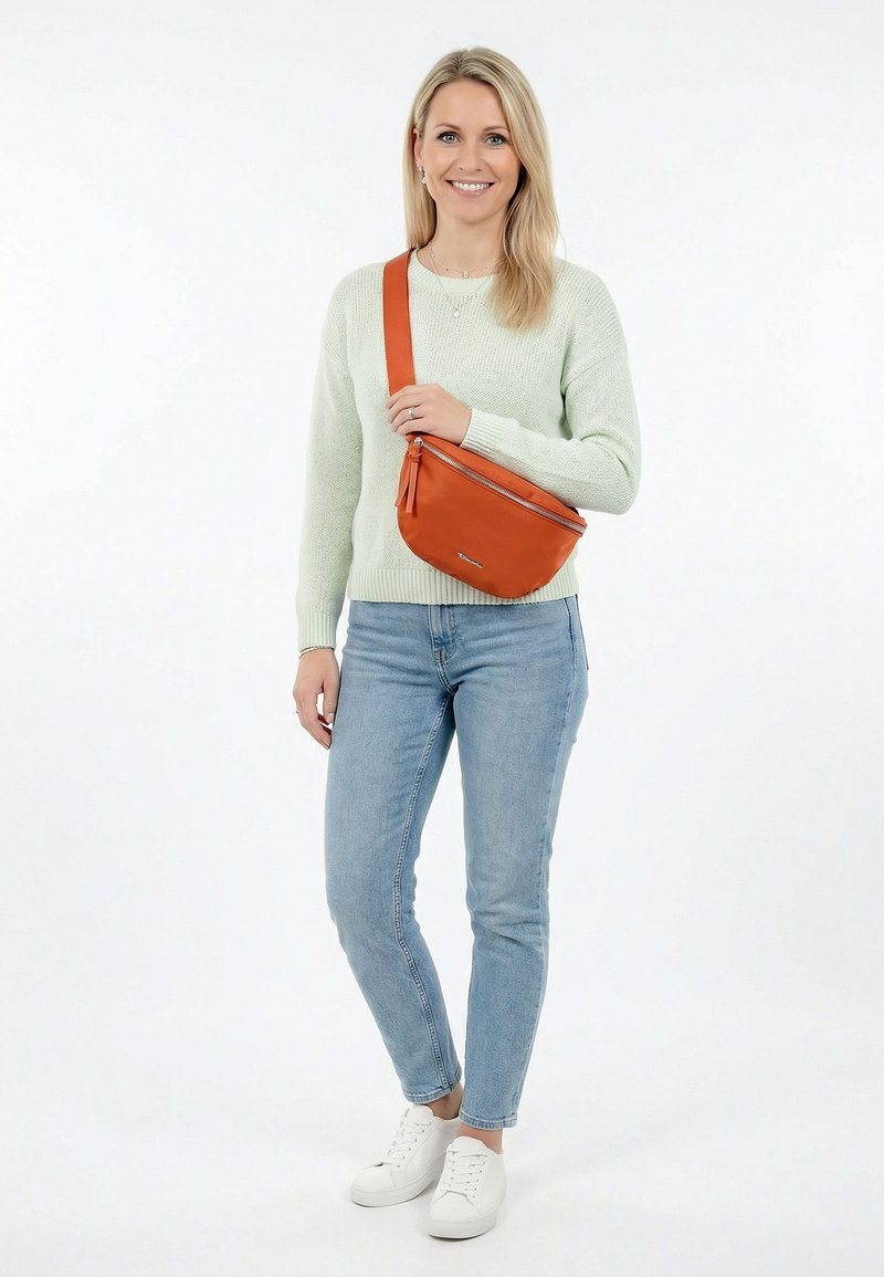 Femme souriante, portant un pull vert clair, un jean bleu, des baskets blanches et un sac orange porté en bandoulière sur sa poitrine, sur fond blanc.