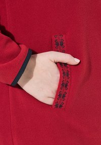 Punainen fleece-takki, jossa on sivutasku, jota koristaa musta hämähäkinseittikirjonta ja kontrastina mustat hihansuut.