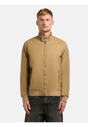 Mężczyzna w beżowej kurtce na zamek z kołnierzem typu stójka i ściągaczami na rękawach, stojący na jednolitym jasnym tle.