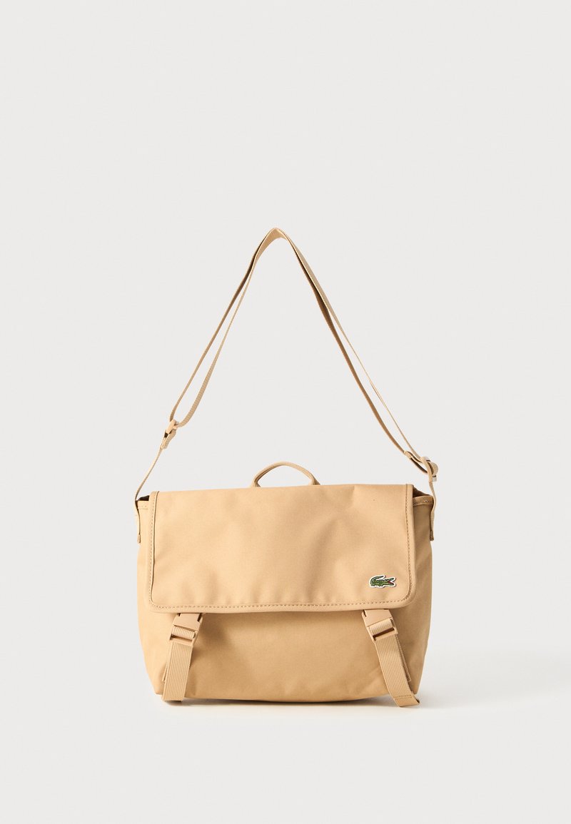 Beige lerret messengerbag med klafflås, justerbar stropp, topphåndtak, og grønn logo-detalj på forsiden. Kompakt rektangulær form.