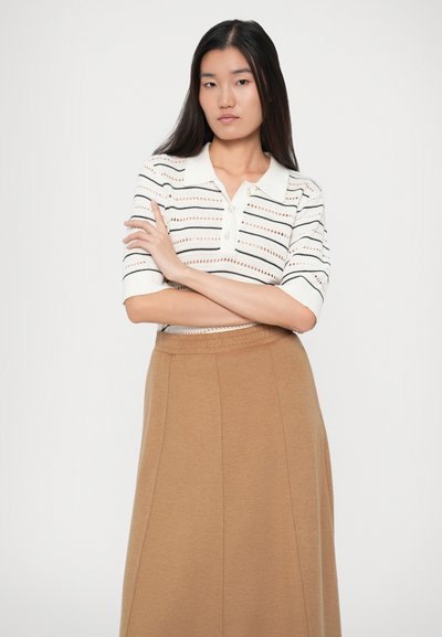 Giovane donna con lunghi capelli scuri che indossa un top a maglia bianco a righe con maniche corte e una gonna a vita alta color cammello, in piedi con le braccia incrociate.