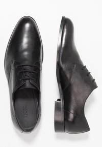 Une paire de chaussures de ville noires en cuir, l'une vue de dessus et l'autre de côté, sur un fond blanc.