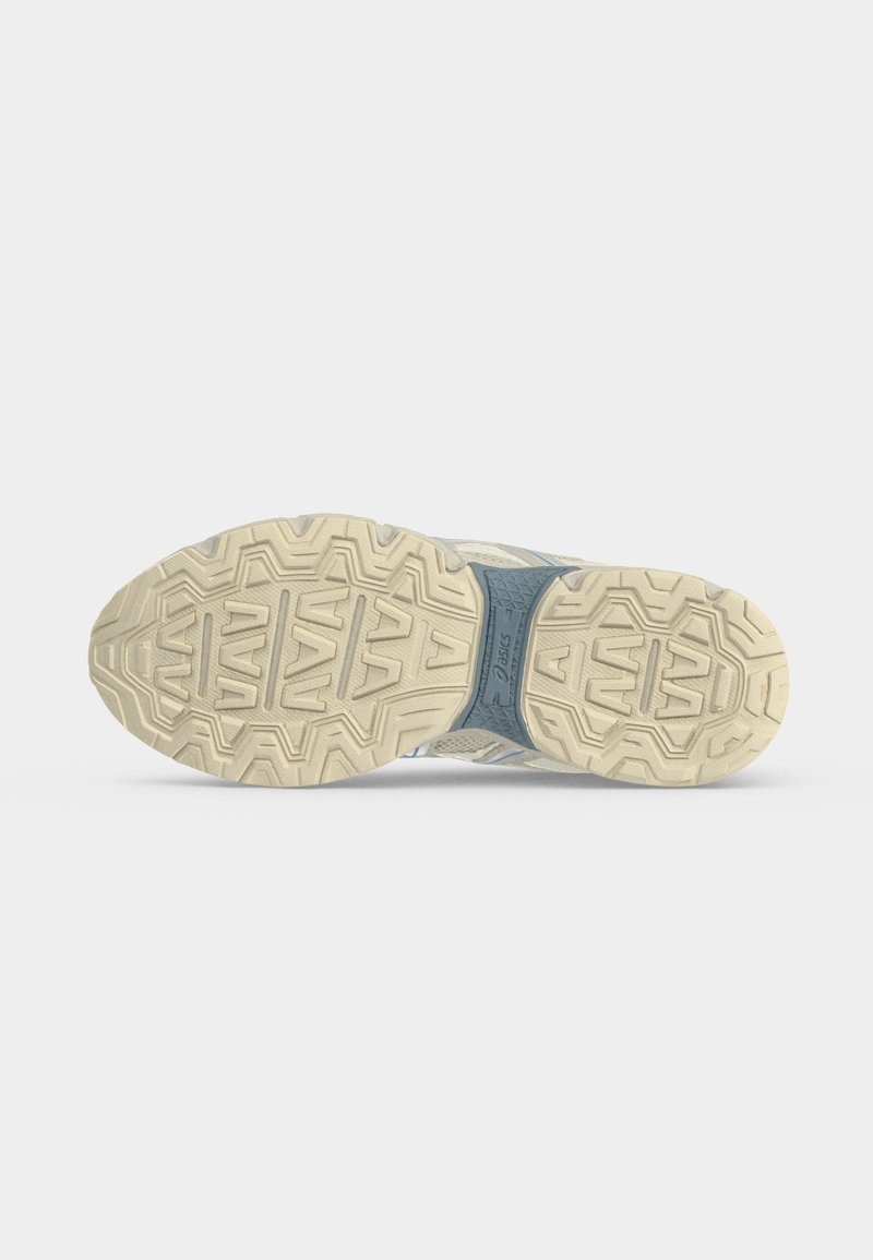 Suela de una zapatilla beige y azul que muestra un patrón de dibujo texturizado y el logo de la marca sobre un fondo liso gris claro.