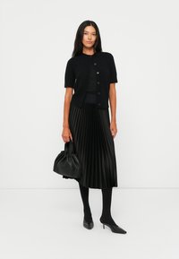 Cardigan nero lavorato a maglia con maniche corte e bottoni, abbinato a una gonna nera a pieghe, collant neri, una borsa nera e scarpe a punta.