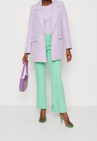 Lila sako přes světle fialový top, doplněné mátovými zelenými kalhotami. Drží lesklou levandulovou kabelku a má na sobě zelené vysoké podpatky se zapínáním na řemínky.