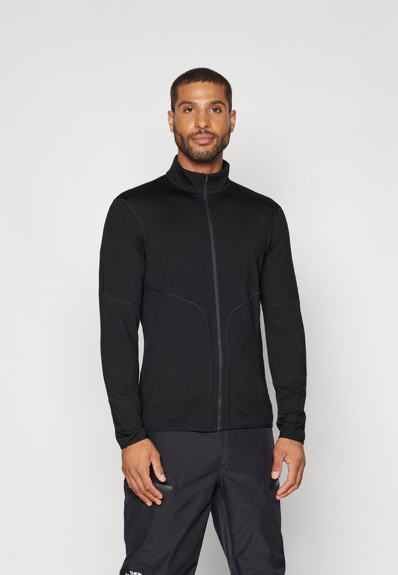 Veste noire à manches longues zippée avec col haut, texture lisse et détails de coutures courbes, accompagnée d'un pantalon sombre.