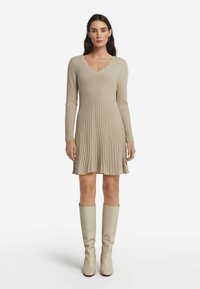 Beige Rippstrickkleid mit V-Ausschnitt, langen Ärmeln und einem ausgestellten Saum. Kombiniert mit kniehohen cremefarbenen Stiefeln. Glatte Textur und taillierte Passform.