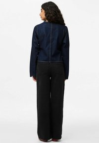 Sieviete stāv, skatoties prom, valkājot tumši zilu džinsa jaku un augstas jostasvietas platajam melnajām džinsu biksēm uz balta fona.