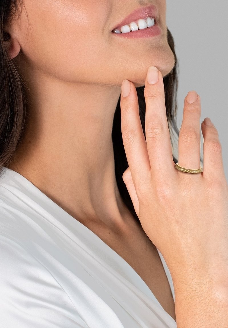 Aranyszínű gyűrű csavarodott mintázattal, az ujjhegyen viselve. Simább textúrájú, fehér ruházat és semleges bőrszín hátterében.