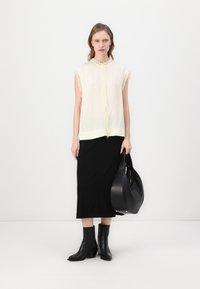 Sleeveless light cream top with fringe trim, paired with a black form-fitting skirt. Model holds a rounded black handbag and wears black ankle boots.