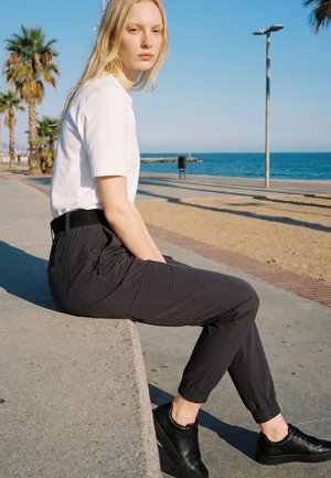 Une femme en tee-shirt blanc, pantalon noir et chaussures noires est assise sur une promenade en bord de mer, avec des palmiers et l’océan derrière elle.