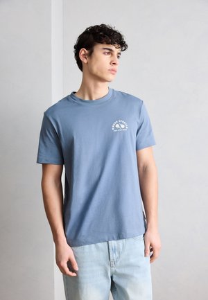 Mladý muž v modrom tričku s nápisom "MOPD SPORTS" a svetlomodrými džínsami, pozerá sa bokom na jednofarebné svetlosivé pozadie.