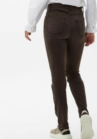 Osoba v tmavě hnědých úzkých kalhotách, bílé blůze s nařasenými manžetami a béžových teniskách kráčí vpřed na bílém pozadí.