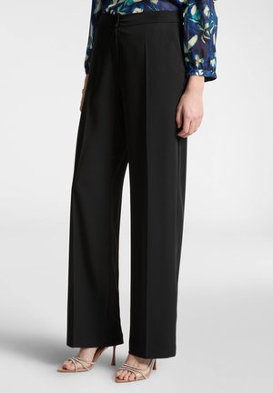 Pantaloni neri a gamba larga con una texture liscia, dotati di pieghe frontali, una cerniera nascosta e abbinati a sandali con tacco a strappo.