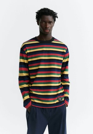 Homme aux cheveux courts et bouclés porte une chemise à manches longues avec des rayures horizontales rouges, jaunes, vertes et bleu marine, les mains dans les poches d’un pantalon sombre.