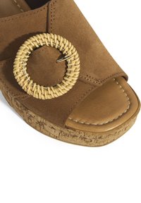 Braune Wildleder-Wedge-Sandale mit weitem Öffnungsbereich und einem runden, geflochtenen Rattan-Accente. Die Sohle weist ein strukturiertes Korkdetail auf.